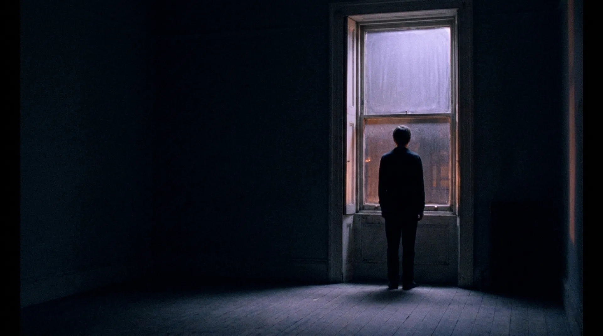 A lone person seen from behind, small in a dark room, standing at a tall window at night and looking out